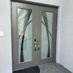 Double gray glass doors with modern decorative patterns enhanced by 3M decorative film, metal handles, and a black semicircular doormat, set against a light-colored wall with a window above.