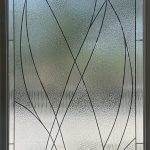 Frosted glass door panel with a geometric pattern of intersecting curved and straight lines, enhanced by decorative film; blurred outdoor background visible through the glass.