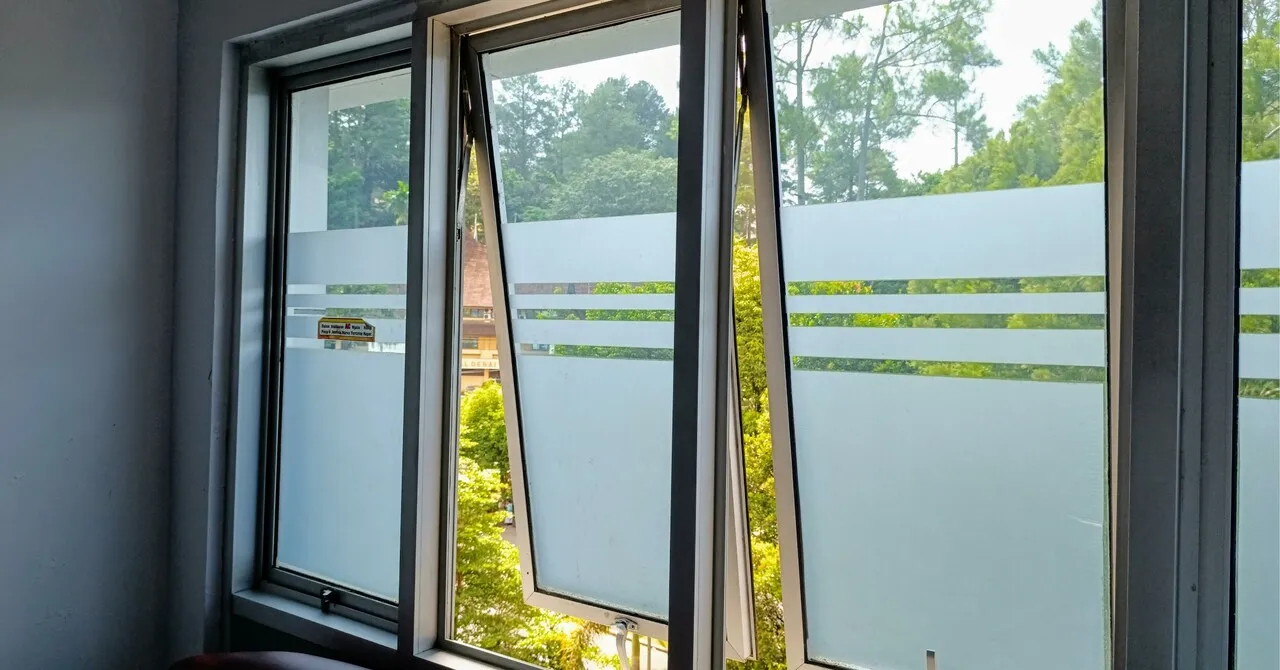 Three glass windows, two of which are slightly open, have frosted lower halves with horizontal clear lines; trees and a building are visible outside in daylight.