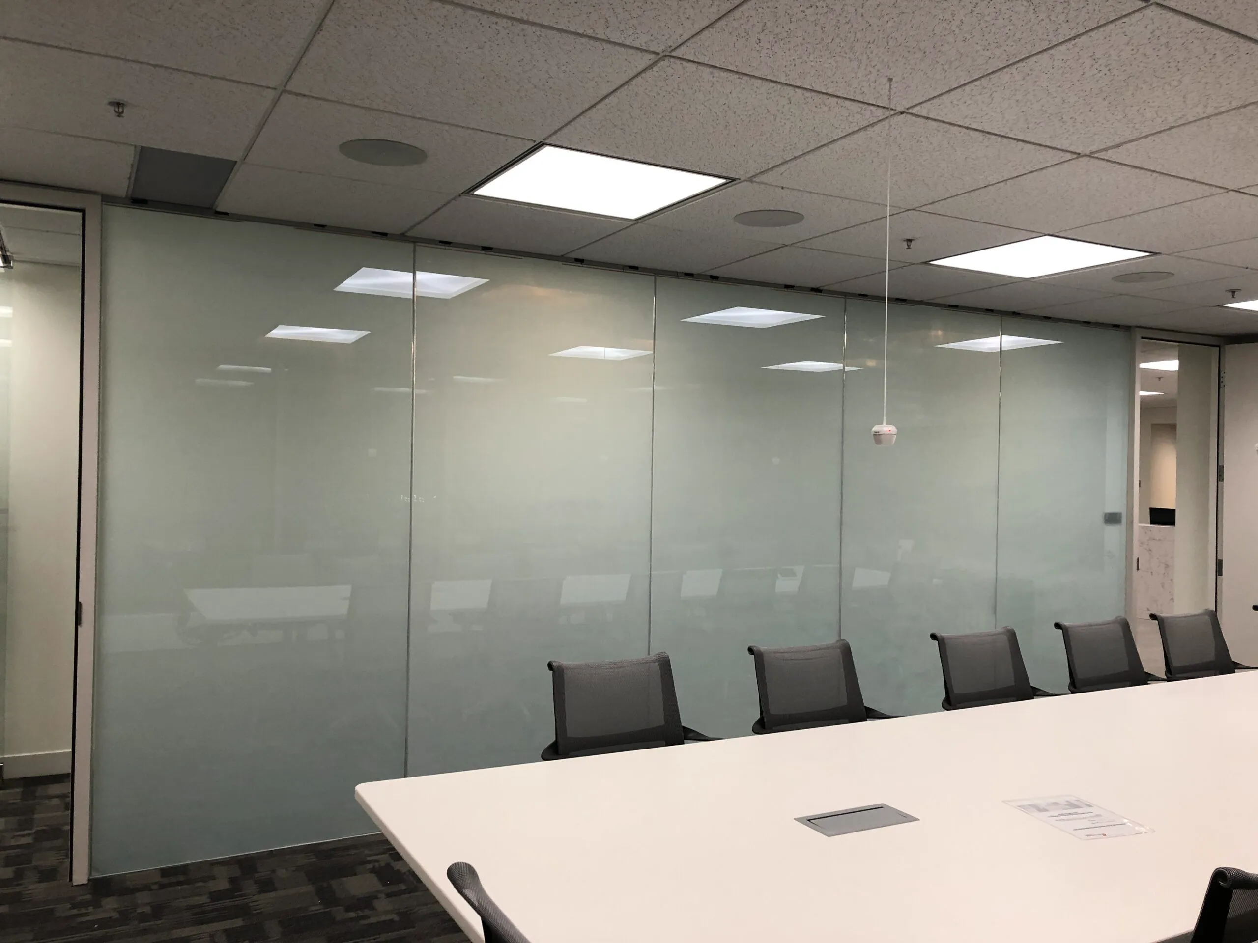A modern conference room with a white table, gray chairs, and frosted glass walls under fluorescent ceiling lights.