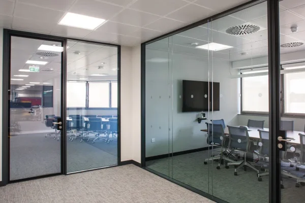 switchable-smart-film-screens-ho Modern office space with glass-walled meeting rooms, gray carpet, ceiling lights, and conference tables with chairs.
