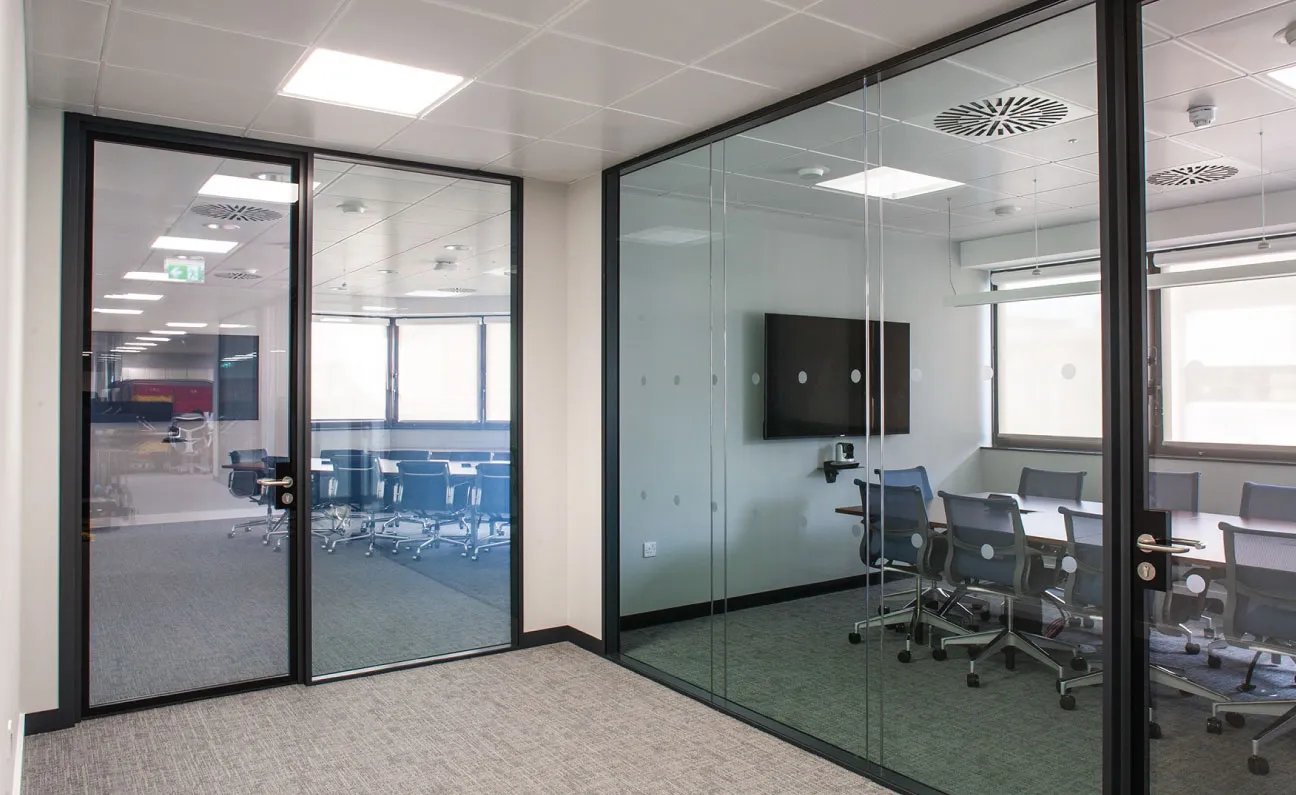 Modern office space with glass-walled meeting rooms, gray carpet, ceiling lights, and conference tables with chairs.