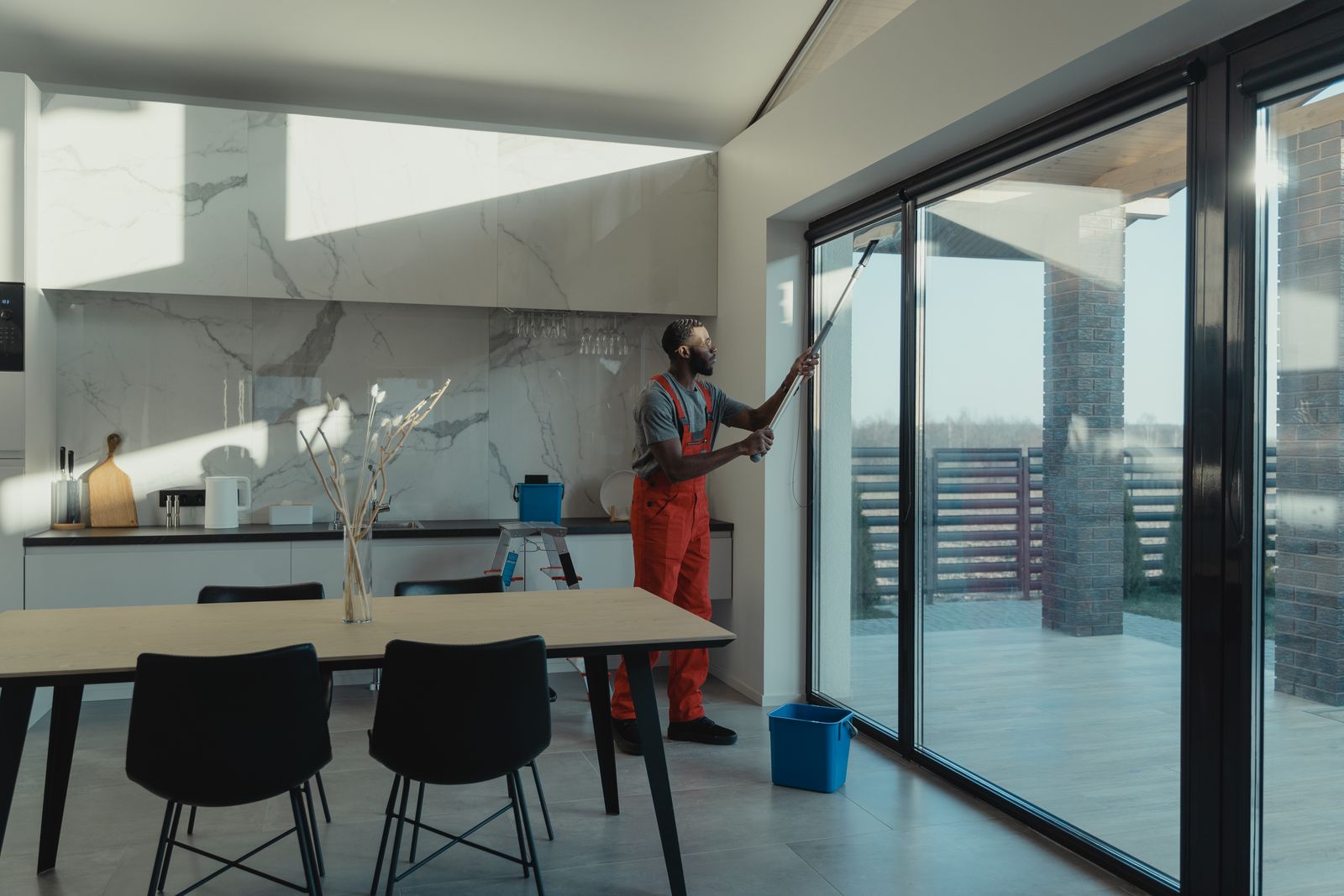 Technician in overalls working on a floor-to-ceiling glass wall in a bright kitchen, the kind of job where people ask can window film be removed cleanly